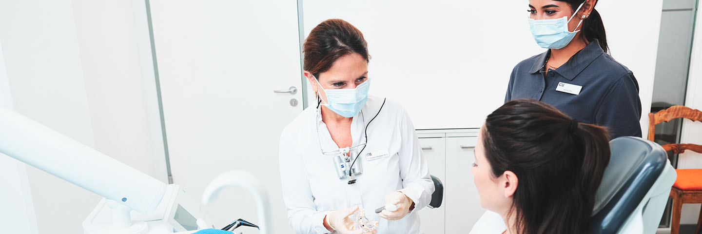 Patientin sitzt im Behandlungszimmer auf dem Behandlungsstuhl und bekommt etwas von der Zahnärztin erklärt, ihre Assistentin steht daneben